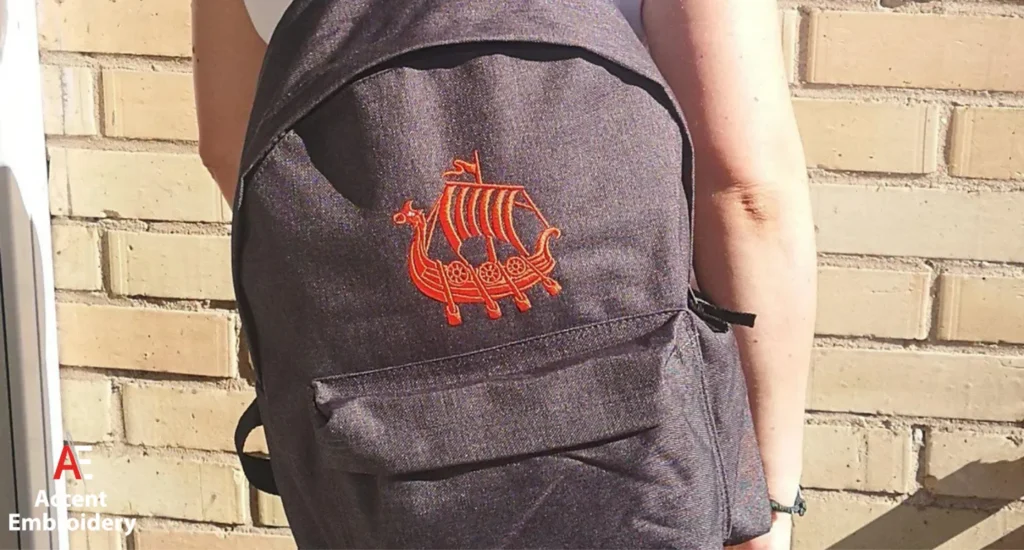 A woman holds a backpack featuring a boat design, showcasing custom embroidery on the fabric. A woman holds a backpack featuring a boat design, showcasing custom embroidery on the fabric.